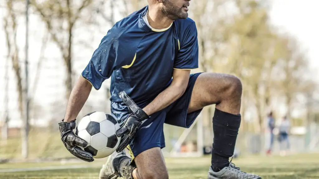 What do goalkeepers wear under gloves - A goalkeeper on his knee and holding a ball
